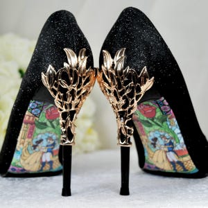 Può includere: Scarpe con tacco alto nere con decorazioni in foglia d'oro. I tacchi presentano un design stampato di Belle e la Bestia del film Disney La Bella e la Bestia.
