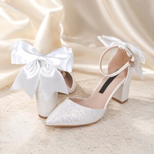 May include: A pair of white glittery block-heel pumps featuring a pointed toe and an ankle strap. Each shoe is adorned with a large, oversized white satin bow attached to the back of the heel, providing a decorative accent to the shimmering footwear.