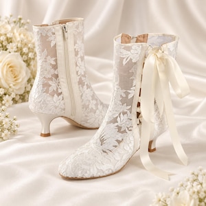 May include: A pair of white ankle boots featuring a sheer mesh overlay with an intricate white floral lace pattern. The boots have a side zipper, a kitten heel, and a decorative satin ribbon tied in a bow at the back of the ankle collar.