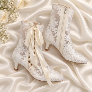 May include: A pair of white lace ankle boots featuring a floral pattern, a kitten heel, and a front ribbon lace-up closure. One boot displays a decorative satin ribbon tied in a bow, while the other shows a side zipper closure detail.