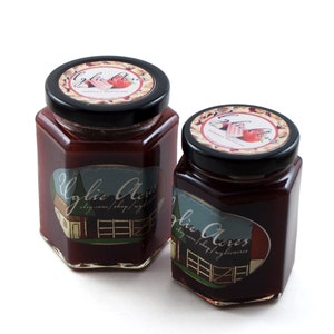 May include: Two jars of strawberry rhubarb jam with black lids. The labels feature the text "Cylie Acres" and a farm scene. The jar lids have a strawberry design. The jars are filled with a red jam.