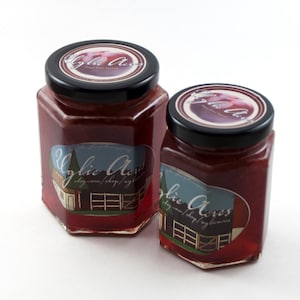 Op de afbeelding: Twee glazen potten met rode vruchtenspread met een wit etiket waarop "Uglie Acres" en een websiteadres staat. De kleinere pot staat voor de grotere.