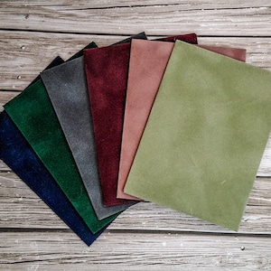 K&ouml;nnte beinhalten: Eine Auswahl an Wildleder-Stoffmustern in verschiedenen Farben, darunter Marineblau, Gr&uuml;n, Grau, Burgunderrot, Rosa und Olivgr&uuml;n. Die Muster sind f&auml;cherf&ouml;rmig auf einer Holzoberfl&auml;che angeordnet und zeigen die Textur und Farbvariationen.