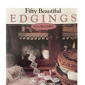 Op de afbeelding: Een boek getiteld "Fifty Beautiful EDGINGS TO CROCHET" met een vintage esthetiek. De omslag toont een close-up van gehaakte kantversiering, garenbollen en een rieten mand.