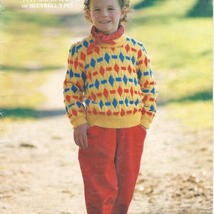 Pode incluir: Uma criança vestindo um suéter amarelo com padrões de losangos vermelhos e azuis, combinado com calças vermelhas e tênis azuis. O suéter tem bainha e punhos canelados. A imagem apresenta o texto "Patons" e "Children's Handknits".