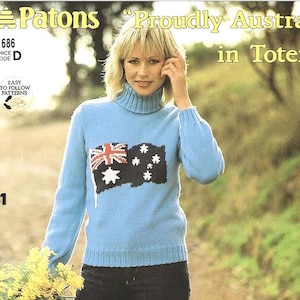 Op de afbeelding: Een blauwe gebreide trui met een wit en rood Australisch vlagontwerp. De trui heeft een rolkraag en lange mouwen. Het model draagt ​​een zwarte jeans en houdt een bos gele bloemen vast.