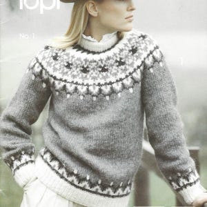 Könnte beinhalten: Eine Frau trägt einen grauen Wollpullover mit einem gemusterten Passe und Bündchen. Der Pullover hat ein weißes und schwarzes geometrisches Design. Das Bild zeigt auch den Text "Alafoss lopi" und "No. 1". Die Frau trägt einen grauen Hut.