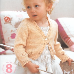 May include: A pale peach knitted cardigan with long sleeves, a V-neckline, and a delicate lace-like border along the cuffs and front edges. The garment features three clear buttons and is presented on a pattern cover titled A Princess Tale.