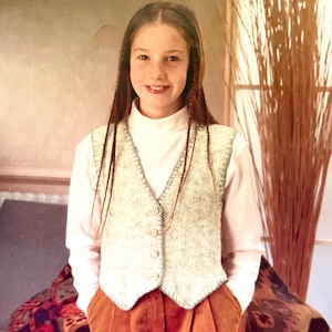 May include: A young person wearing a white turtleneck shirt with a crocheted vest. The vest is white with a light blue trim and has a button closure. The person is wearing brown corduroy trousers and has long brown hair.