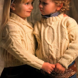 May include: Two children in cream cable-knit sweaters with textured, braided patterns. One child has long blonde hair and a patterned headband. The other has curly blonde hair. They are holding hands. The sweaters are a warm, off-white colour.