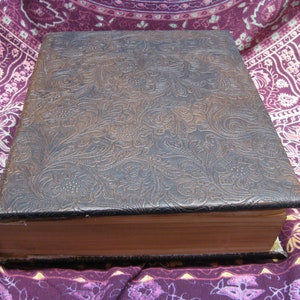 Puede incluir: Un libro encuadernado en cuero marr&oacute;n con un patr&oacute;n floral en relieve en la portada. El libro est&aacute; cerrado y sobre una tela con estampado morado y blanco.