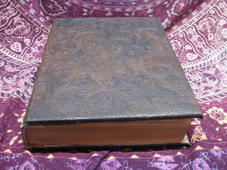 Puede incluir: Un libro encuadernado en cuero marr&oacute;n con un patr&oacute;n floral en relieve en la portada. El libro est&aacute; cerrado y sobre una tela con estampado morado y blanco.