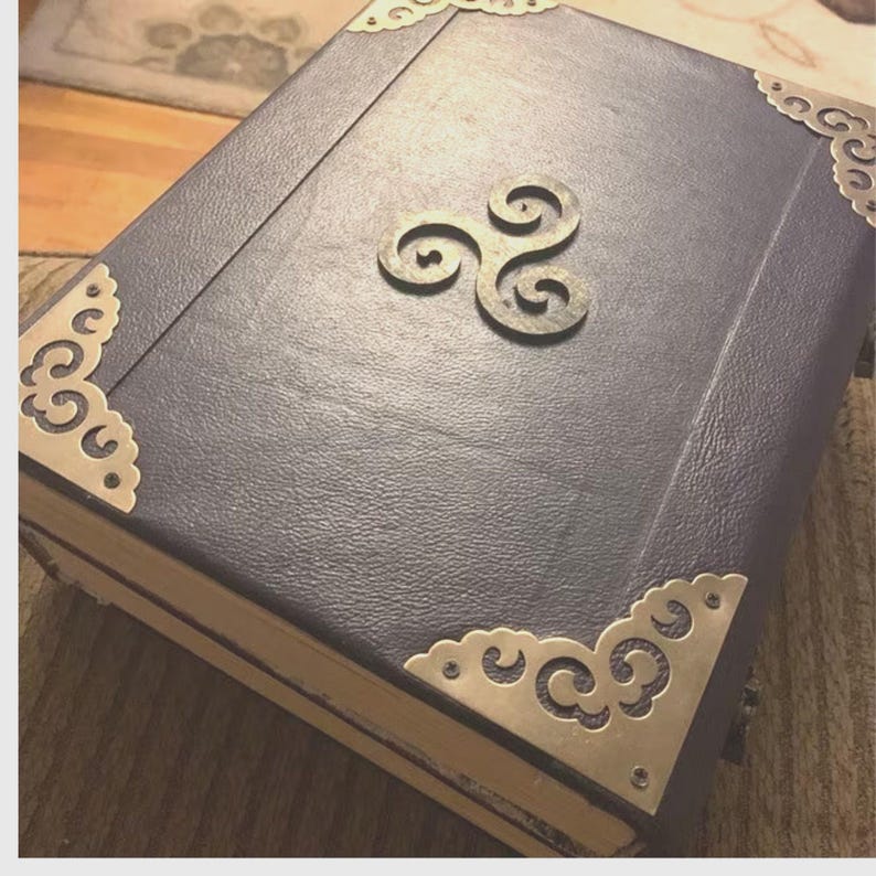 Pu&ograve; includere: Un libro rilegato in pelle blu scuro con protezioni angolari in ottone ornate e un emblema decorativo centrale. I bordi del libro sono invecchiati e la copertina ha un aspetto strutturato, che suggerisce uno stile vintage o antico.