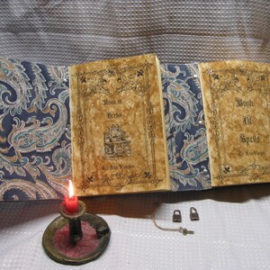 May include: Two antique-looking books with paisley covers. The books are titled "Book of Herbs" and "Book of Spells". A lit candle and two padlocks are on a white surface.