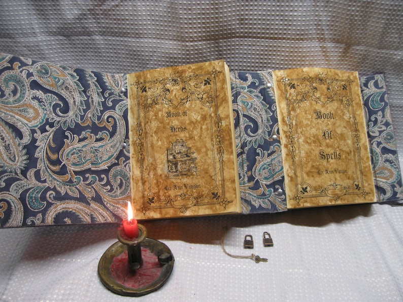 May include: Two antique-looking books with paisley covers. The books are titled "Book of Herbs" and "Book of Spells". A lit candle and two padlocks are on a white surface.