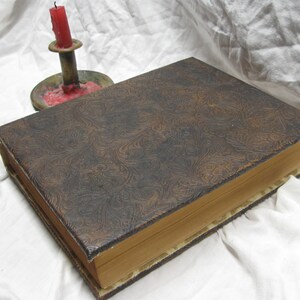 Puede incluir: Un libro encuadernado en cuero marr&oacute;n con un patr&oacute;n decorativo en relieve en la portada. El libro est&aacute; cerrado y se encuentra sobre una superficie blanca.