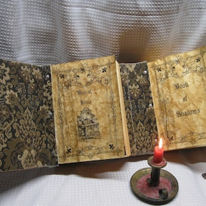 May include: An open book with two pages titled "Book of Herbs" and "Book of Shadows". The book has a brown and gold patterned cover and is sitting on a white surface with a lit candle and two keys.