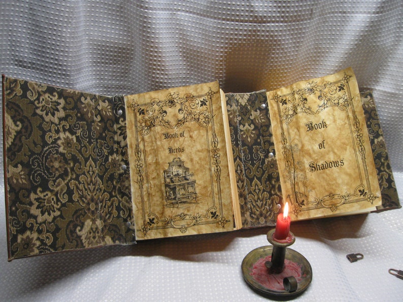 May include: An open book with two pages titled "Book of Herbs" and "Book of Shadows". The book has a brown and gold patterned cover and is sitting on a white surface with a lit candle and two keys.