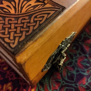 Pu&ograve; includere: Primo piano di un diario rilegato in pelle fatto a mano. La copertina presenta intricati disegni di nodi celtici e floreali in marrone scuro e oro. Un fermaglio decorativo in metallo fissa il bordo del diario.