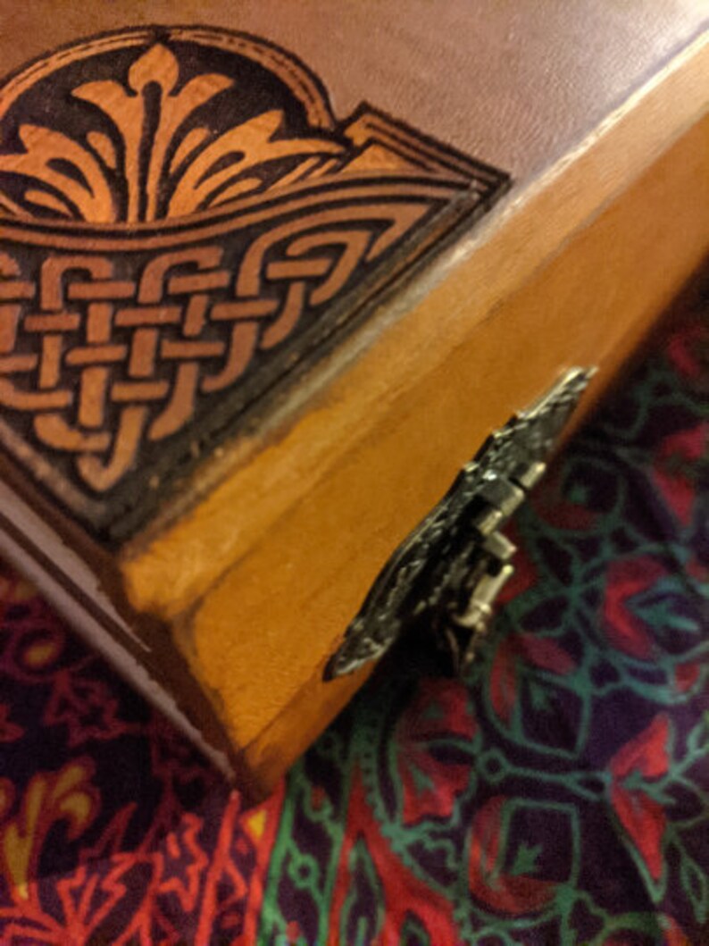 Pu&ograve; includere: Primo piano di un diario rilegato in pelle fatto a mano. La copertina presenta intricati disegni di nodi celtici e floreali in marrone scuro e oro. Un fermaglio decorativo in metallo fissa il bordo del diario.