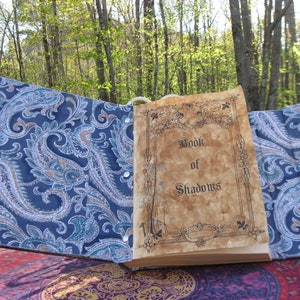 Może przedstawiać: Otwarta książka z okładką w wz&oacute;r paisley. Książka nosi tytuł "Book of Shadows" i jest otwarta na pustej stronie.
