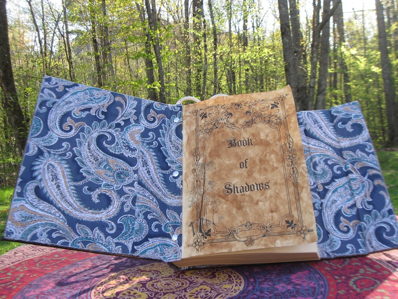 Może przedstawiać: Otwarta książka z okładką w wz&oacute;r paisley. Książka nosi tytuł "Book of Shadows" i jest otwarta na pustej stronie.