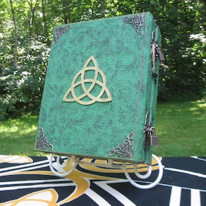 Op de afbeelding: Een groen lederen gebonden dagboek met een gouden triquetra-symbool op de voorzijde. Het dagboek heeft twee sloten en staat op een witte standaard.