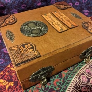 Pu&ograve; includere: Una scatola in pelle marrone con dettagli metallici decorativi e disegni angolari. La scatola presenta un emblema metallico dell'albero della vita e due piccole etichette rettangolari con la scritta "Book of Shadows" e "Book of Herbs". Una chiusura in metallo chiude la scatola.