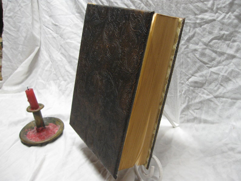 Puede incluir: Un libro encuadernado en cuero marr&oacute;n oscuro con un patr&oacute;n decorativo en relieve. El libro tiene un lomo grueso y est&aacute; cerrado.