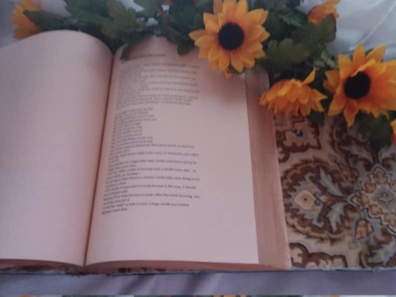 Pu&ograve; includere: Un libro aperto con testo sulla pagina destra, con una composizione floreale di girasoli e verde in alto. Il libro poggia su una superficie di tessuto fantasia.