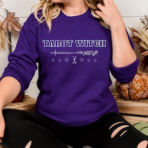 Puede incluir: Una sudadera morada con el texto "Tarot Witch" en letras blancas. El diseño incluye una espada, una copa, un pentagrama y una varita.
