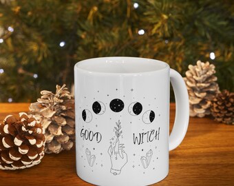 May include: White ceramic mug with a black and white design of a hand holding a sprig of leaves, surrounded by moons and stars. The text "GOOD WITCH" is printed on the mug.