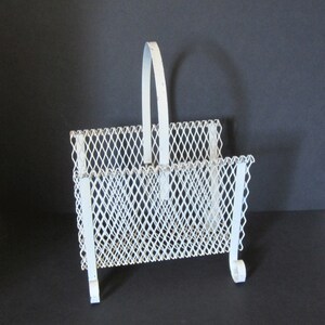 Könnte beinhalten: Ein weißer Zeitschriftenständer aus Metall mit Griff. Der Ständer hat ein rautenförmiges Maschendesign und einen gebogenen Fuß. Der Griff ist gewölbt und der Ständer ist weiß lackiert. Er ist für Zeitschriften oder andere Lesematerialien konzipiert.