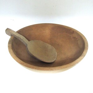 May include: A round, light brown wooden bowl with a smooth interior surface and a matching wooden spoon resting inside. The spoon features a long, flat handle and a wide, oval-shaped bowl, both showing a natural wood grain and light brown finish.