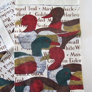 May include: White fabric with a duck print. The ducks have red, brown, green, and yellow details. The fabric also has text with duck names like "Black Duck" and "Mallard".