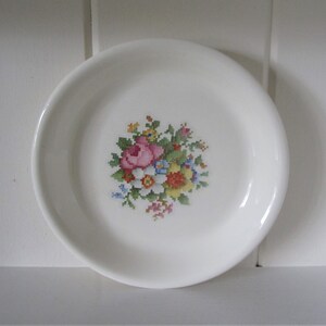 Könnte beinhalten: Ein weißer Keramikteller mit einem floralen Kreuzstichmuster in der Mitte. Das Design zeigt rosa Rosen, gelbe und blaue Blumen und grüne Blätter. Der Teller hat einen leicht gebogenen Rand.