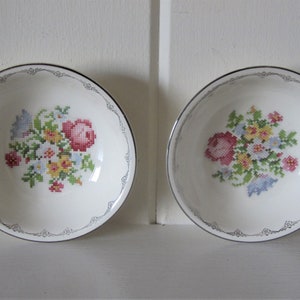 May include: Two white ceramic bowls with a floral cross-stitch design. Each bowl features a central bouquet of pink, red, yellow, and blue flowers. The rims have a silver trim with a decorative pattern.