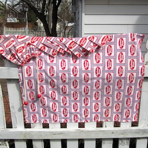 Op de afbeelding: Een wit houten hek met een rood-witte stoffen tafelkleed dat eraan hangt. Het tafelkleed heeft een herhalend patroon van rode vierkanten met de woorden "Strawberry Bread" in wit gedrukt.