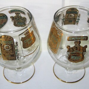 May include: Two clear brandy snifters with gold rims and decorative labels. The labels feature gold and teal accents with text including "Smirnoff Vodka" and "Canada Dry Quinine Water".