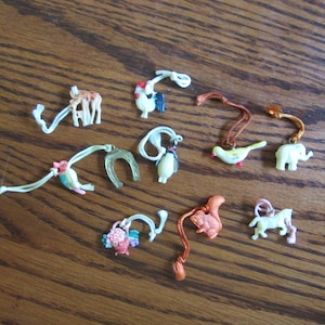 May include: A collection of small, colorful animal charms and a horseshoe charm on a wooden surface. The charms include a giraffe, rooster, bird, elephant, squirrel, horse, and a penguin. Each charm is attached to a white or brown string.