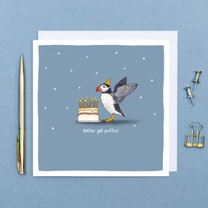 Pu&ograve; includere: Biglietto di auguri con un pulcinella di mare dei cartoni animati con un cappello da festa, accanto a una torta con candele accese. Il biglietto ha il testo "Better get puffin!" su uno sfondo blu con stelle bianche.