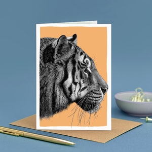 May include: Greeting card featuring a detailed black and white illustration of a tiger's profile against an orange background. The card is on a brown envelope, with a gold pen and small bowl in the background.