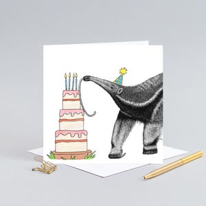 May include: A birthday card featuring a whimsical illustration of an anteater wearing a party hat, reaching for a multi-tiered cake with lit candles. The card is white with a gold pen and paperclip in the background.