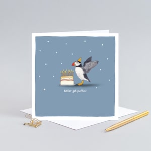 Pu&ograve; includere: Biglietto di auguri con un pulcinella di mare dei cartoni animati con un cappello da festa, accanto a una torta con candele accese. Il biglietto dice "Better get puffin!" su uno sfondo blu con stelle bianche.