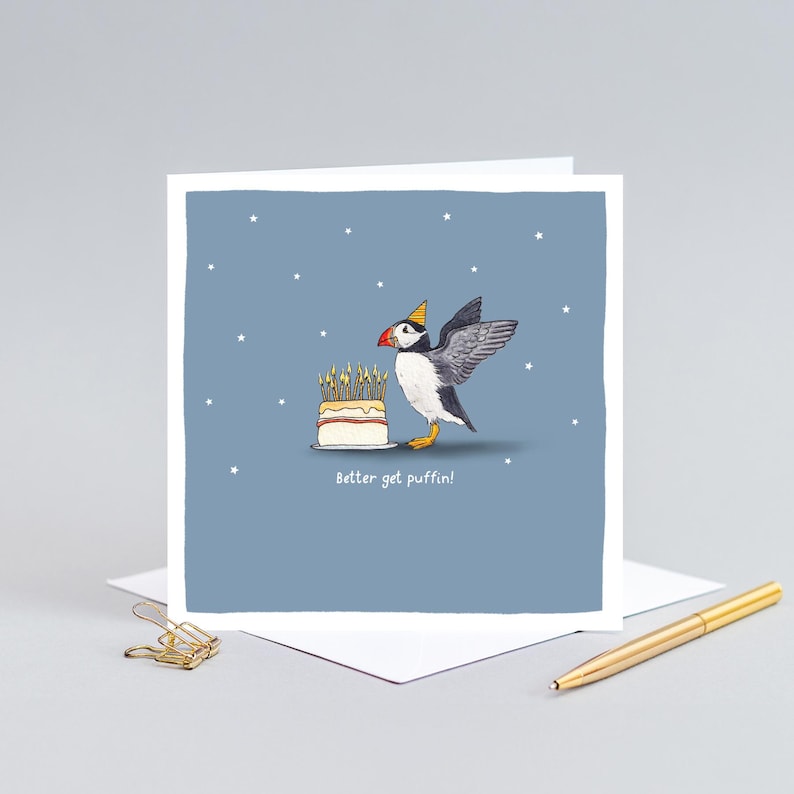 Pu&ograve; includere: Biglietto di auguri con un pulcinella di mare dei cartoni animati con un cappello da festa, accanto a una torta con candele accese. Il biglietto dice "Better get puffin!" su uno sfondo blu con stelle bianche.