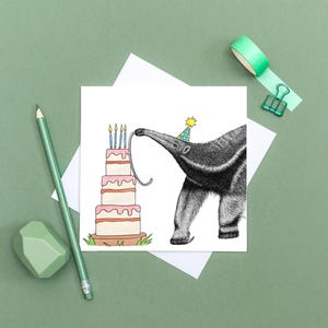 May include: A birthday card featuring a detailed illustration of an anteater wearing a party hat, reaching for the candles on a tiered cake. The cake has pink frosting and is on a wooden base. The card is on a white background with a green pencil and eraser.