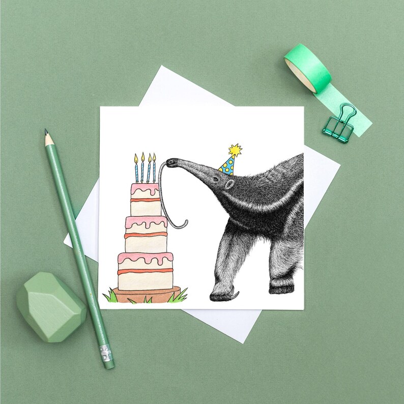 May include: A birthday card featuring a detailed illustration of an anteater wearing a party hat, reaching for the candles on a tiered cake. The cake has pink frosting and is on a wooden base. The card is on a white background with a green pencil and eraser.