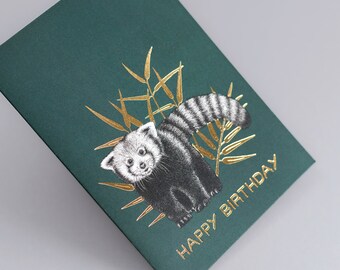 May include: A green card with gold foil accents featuring a red panda with a black and white striped tail. The red panda is surrounded by gold foil leaves. The text "Happy Birthday" is written in gold foil on the card.