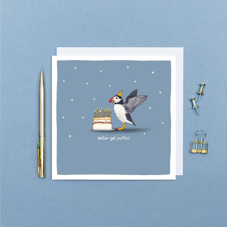 Pu&ograve; includere: Biglietto di auguri con un pulcinella di mare dei cartoni animati con un cappello da festa, accanto a una torta con candele accese. Il biglietto ha il testo "Better get puffin!" su uno sfondo blu con stelle bianche.