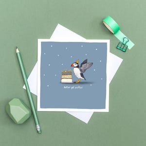 Pu&ograve; includere: Biglietto di auguri quadrato con un pulcinella di mare dei cartoni animati con un cappello da festa, accanto a una torta con candele accese. Il pulcinella di mare ha un'ala tesa. Il testo "Better get puffin!" &egrave; sotto la torta. Lo sfondo &egrave; blu con stelle bianche.